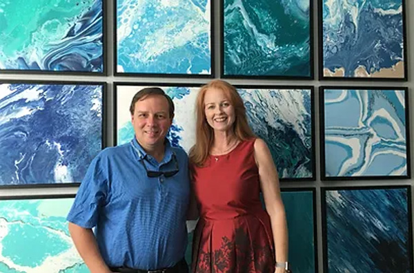 The image features a man and a woman posing in front of an art gallery wall adorned with multiple abstract paintings.