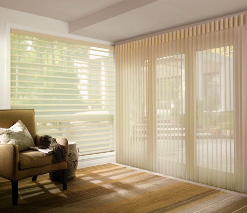 An image of a modern living room interior, featuring a large window with vertical blinds partially drawn, allowing natural light to enter, and a view of the outside through the glass. The room has neutral-toned flooring and walls, with a comfortable sofa in the foreground and a sliding door or patio access point on the right side.