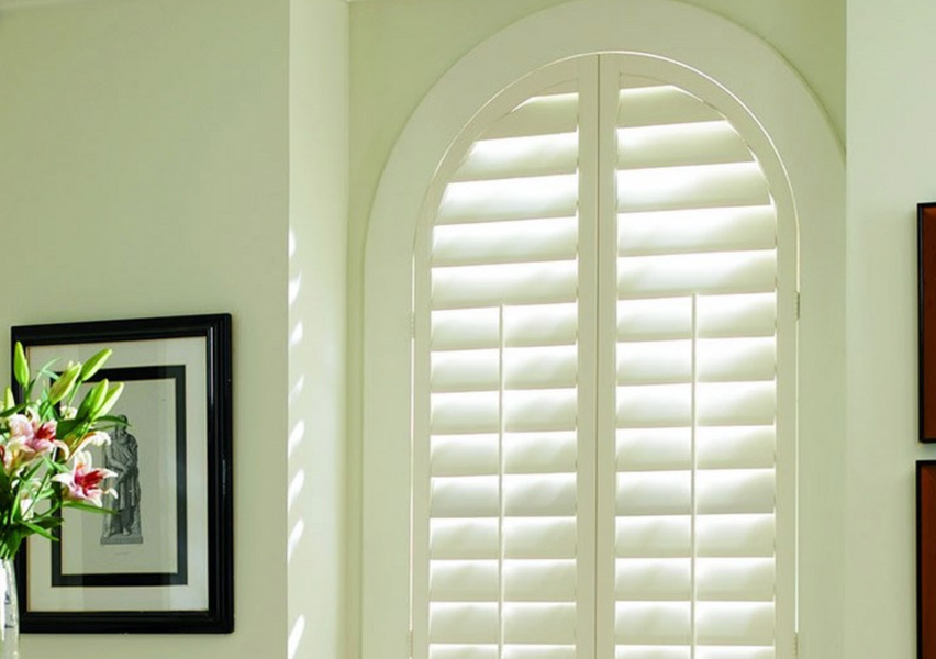 The image shows an interior space with a large arched window covered by white shutters, allowing natural light to enter the room.