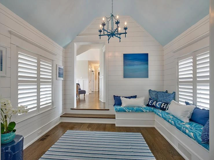 An interior view of a room with white walls, blue accents, and a large window. The room features a cozy seating area with a bench and pillows, a chandelier hanging from the ceiling, and a nautical-themed decor.