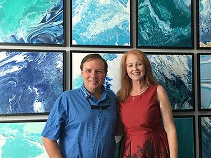 The image shows a man and a woman standing in front of a gallery wall displaying various abstract artworks.